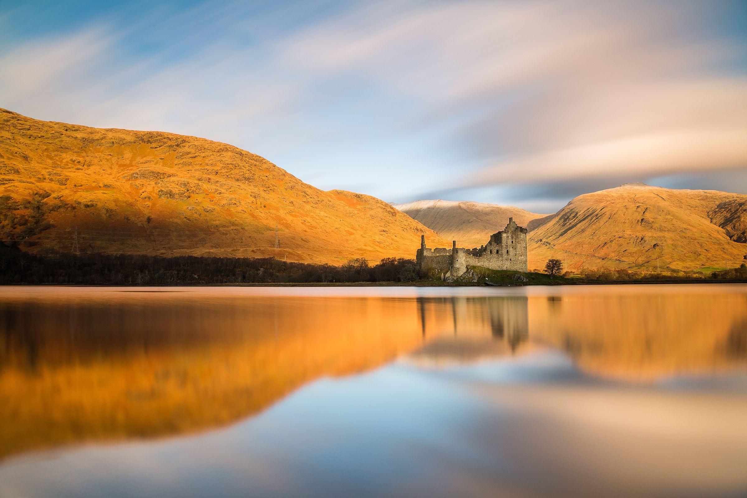Kilchurn Castle at sunrise, its ruin reflected in a still Loch Awe beneath golden hillsides